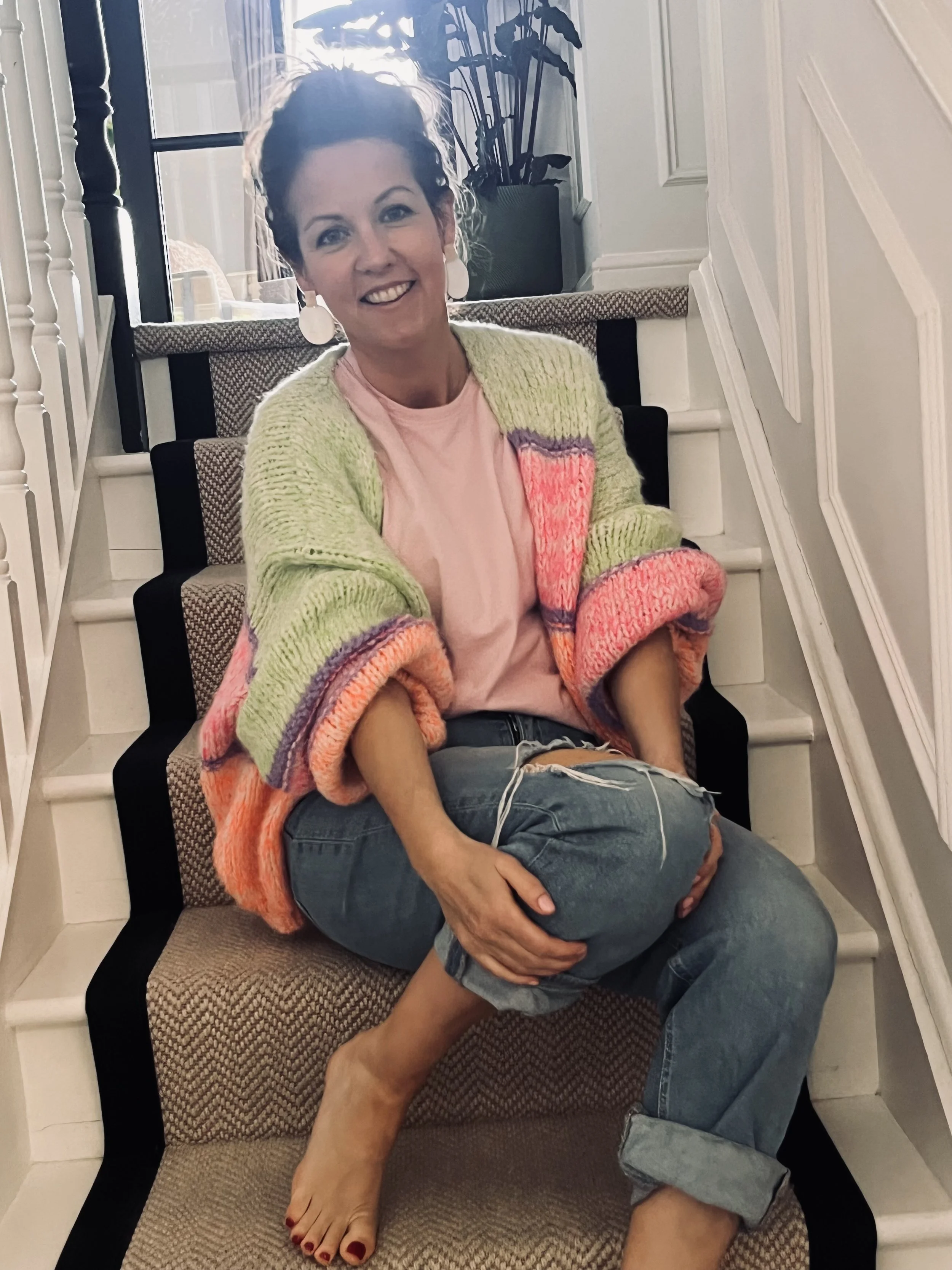 A woman sitting on a staircase with another person lying on her lap, both smiling. The woman wears a colorful knitted sweater, a pink shirt, and earrings. The person in her lap wears ripped jeans and has bare feet.
