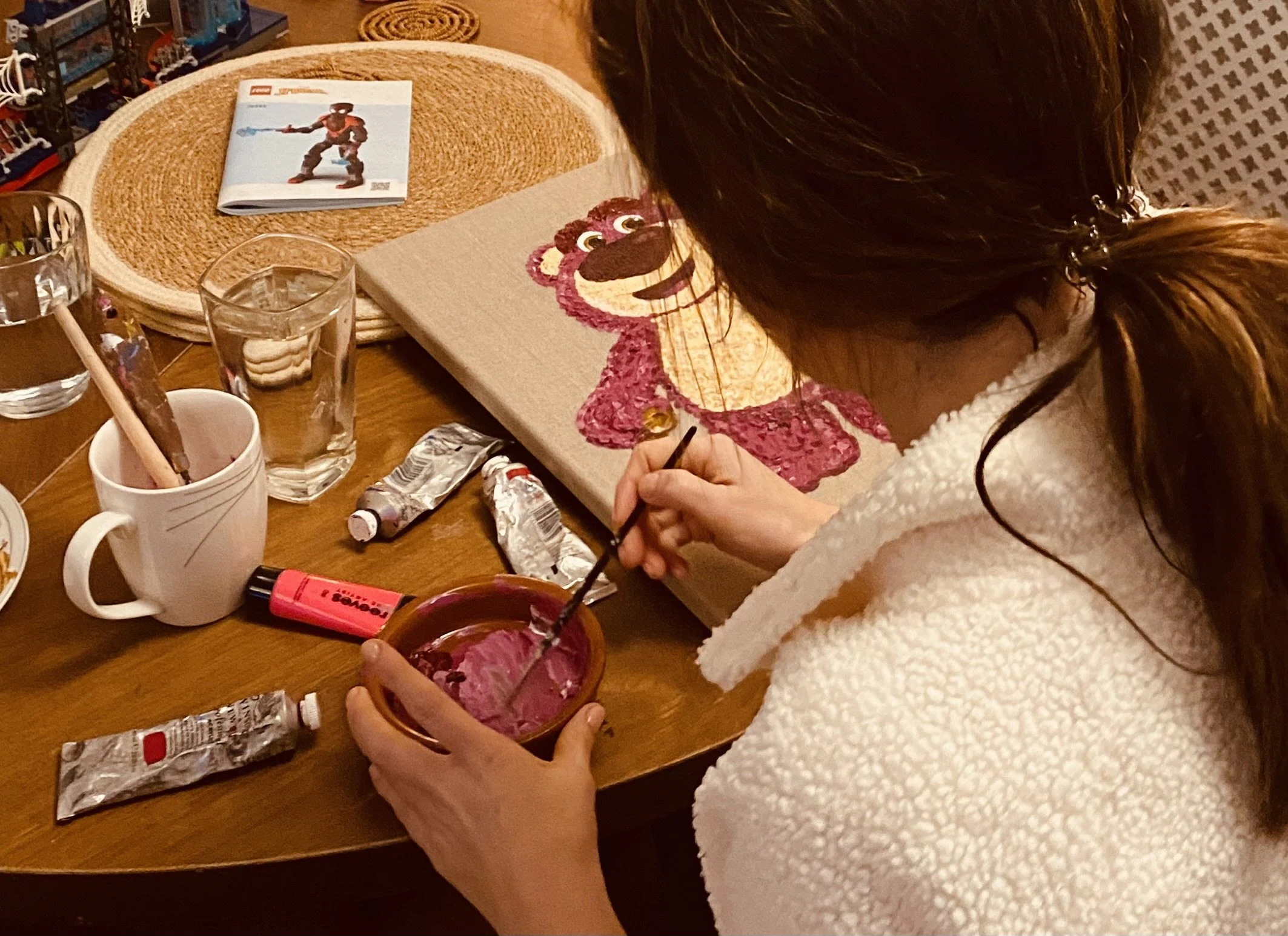 A woman with brown hair tied in a ponytail is painting a lion's face on a canvas. The painting is detailed and colorful. There are art supplies, including tubes of paint, brushes, and water glasses, on the table around her. A LEGO instruction booklet is also on the table.