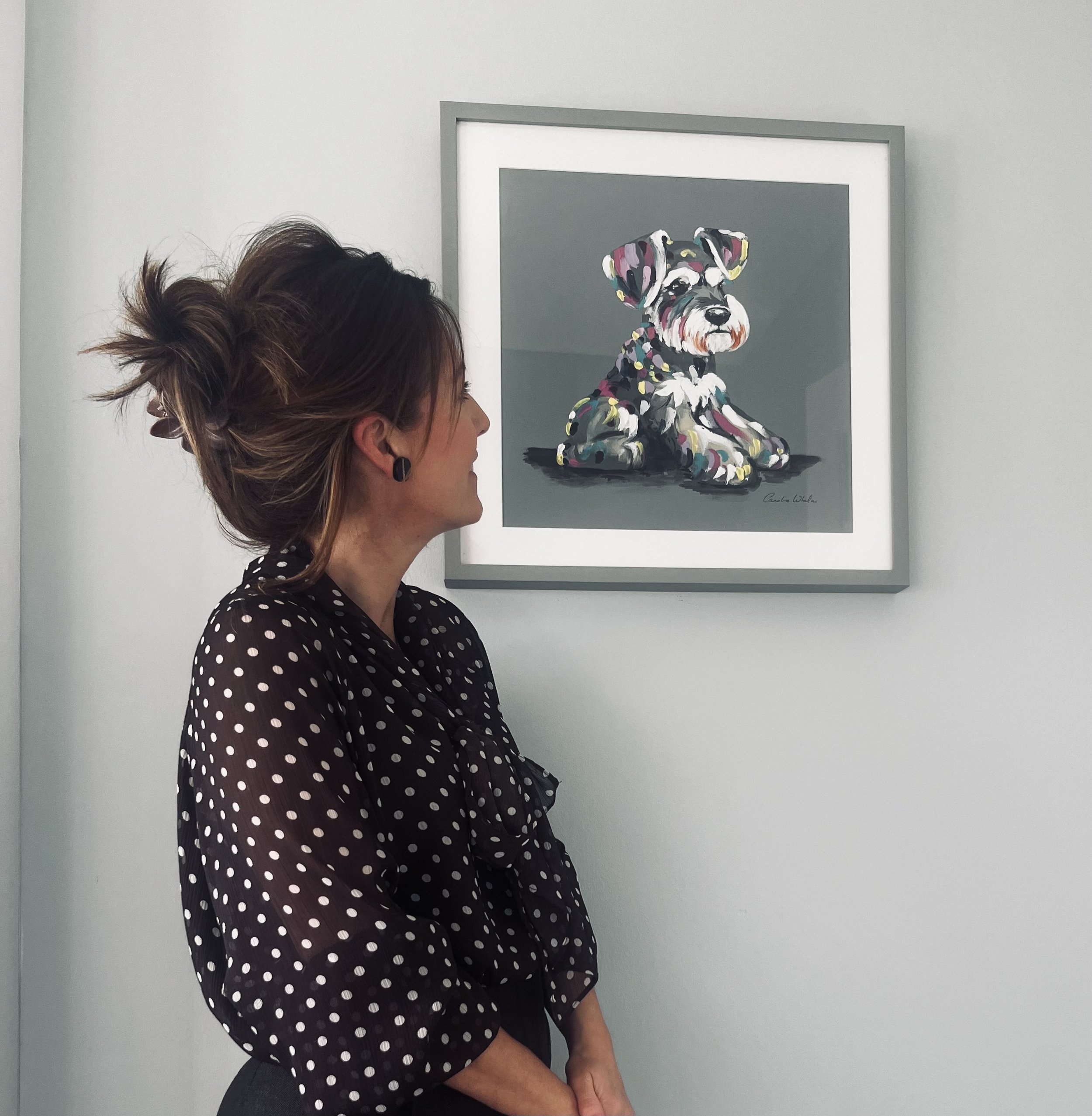 A woman with glasses and earrings wearing a black polka dot blouse, standing in profile in front of a framed painting of a colorful dog on a gray background.