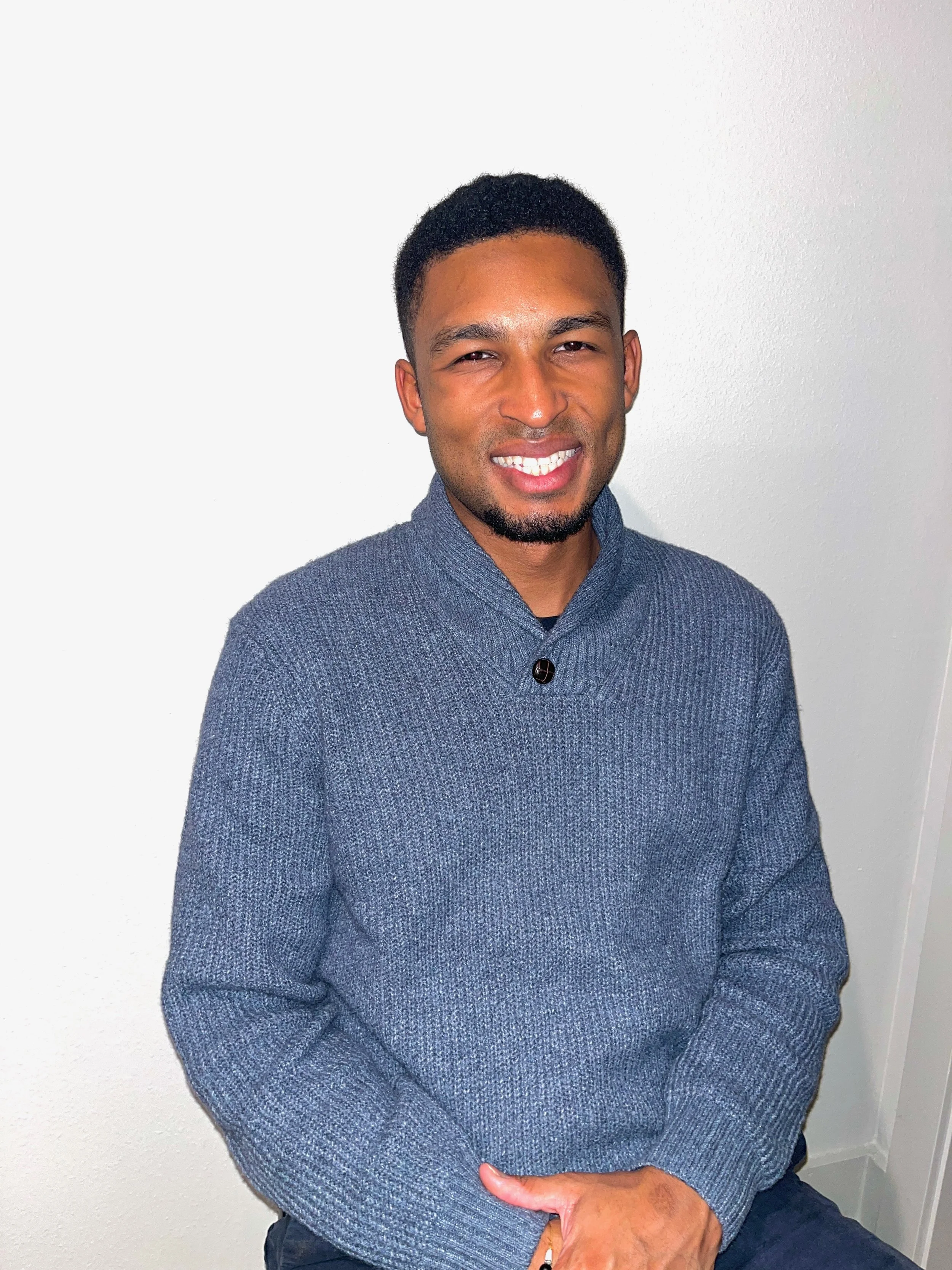 A young man with short black hair and facial hair, wearing a gray textured sweater, sitting against a plain white wall, smiling at the camera.