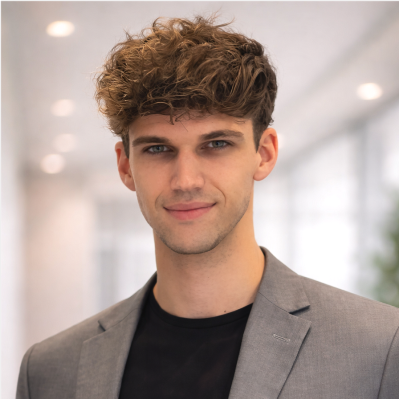 Portrait of a young man with curly brown hair, blue eyes, wearing a gray blazer and black shirt, smiling softly, in a bright office setting.