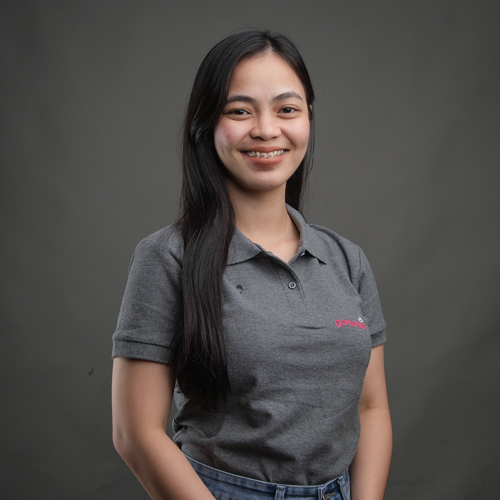 A young woman with long black hair, wearing a gray polo shirt and jeans, smiling in front of a plain dark gray background.