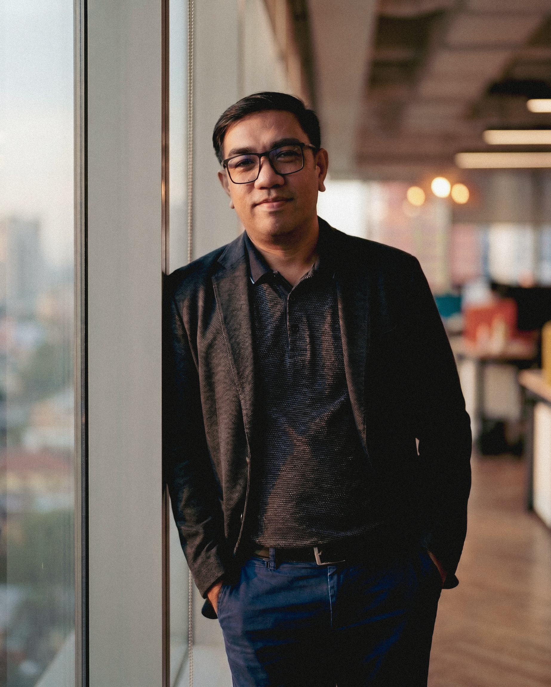 A young man with glasses leaning against a glass wall, smiling, wearing a dark blazer, a patterned shirt, and blue pants in an indoor setting with warm lighting.