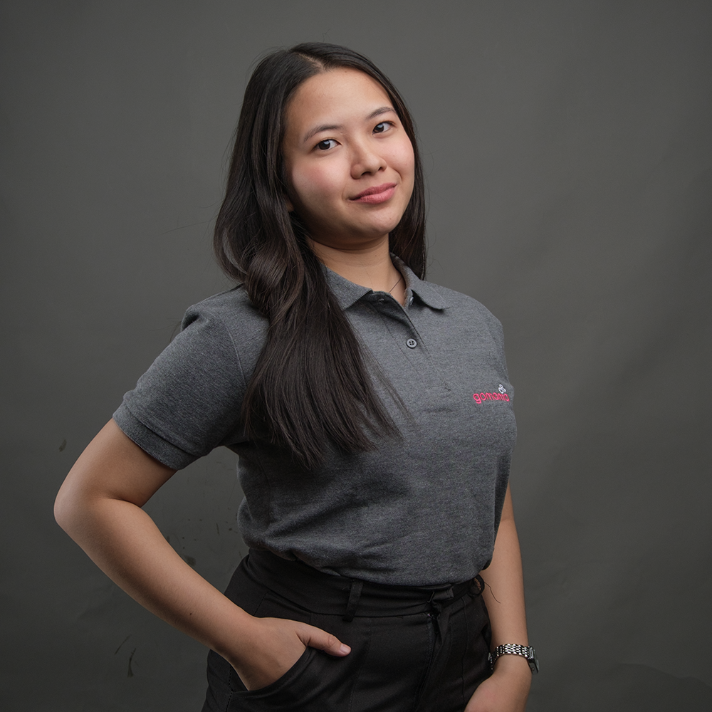 A young woman with long dark hair, wearing a gray polo shirt and black pants, standing in front of a plain gray background with her hand in her pocket, looking at the camera and smiling slightly.