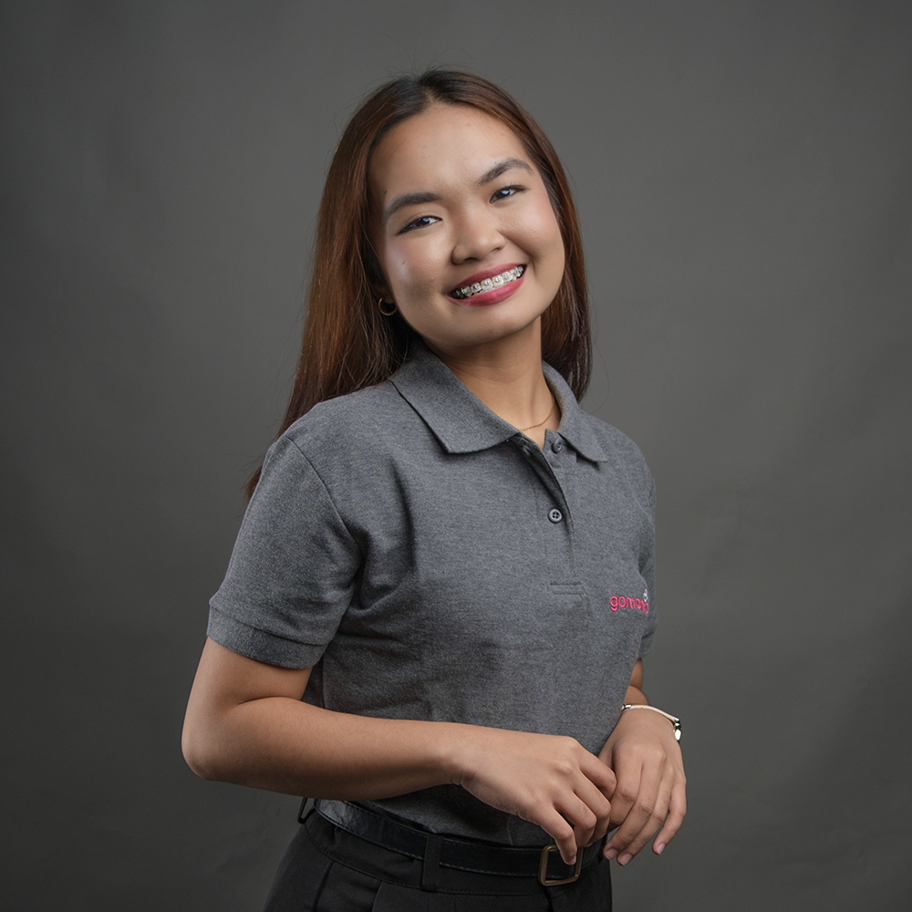 A young woman smiling with braces, wearing a gray polo shirt and black pants, against a gray background.