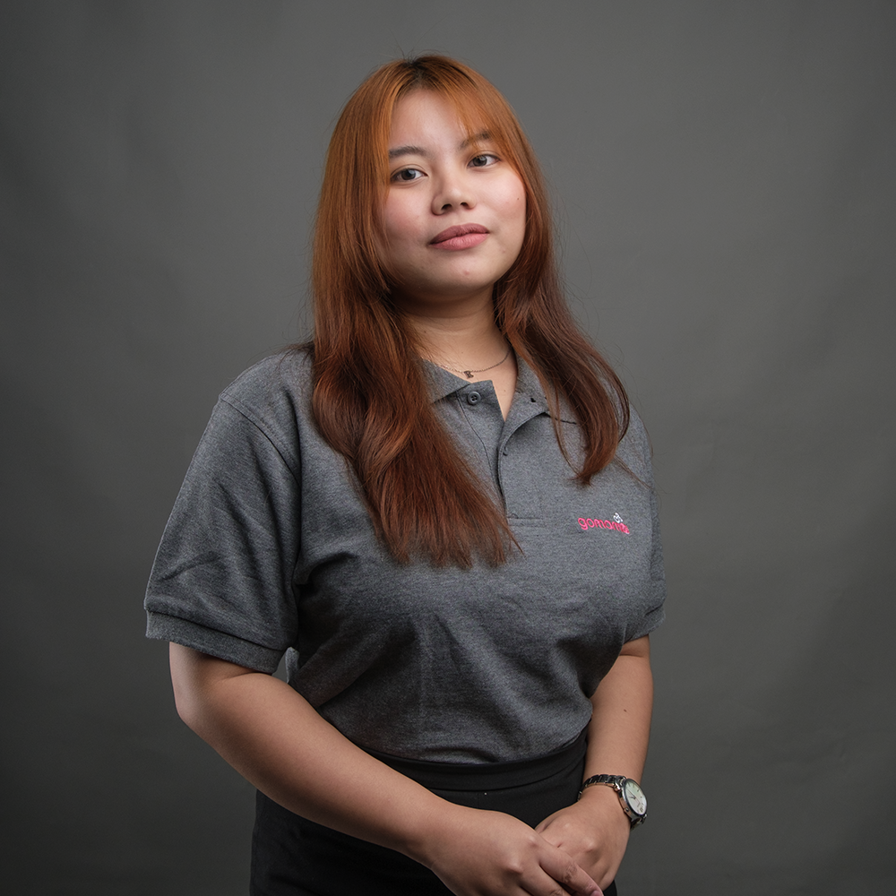 A young woman with long, reddish-brown hair, wearing a gray polo shirt with pink embroidery, standing against a plain gray background.