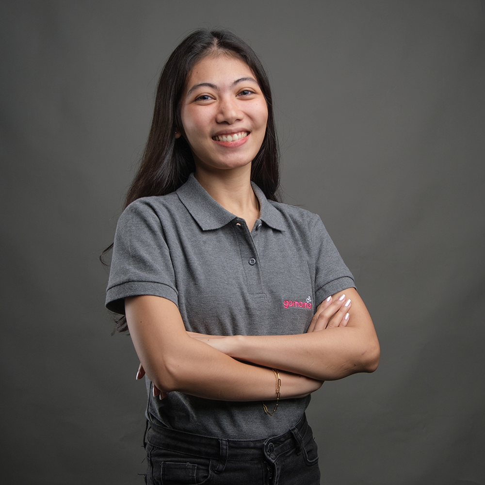 A young woman with long dark hair smiling and crossing her arms, wearing a gray polo shirt, standing against a plain gray background.