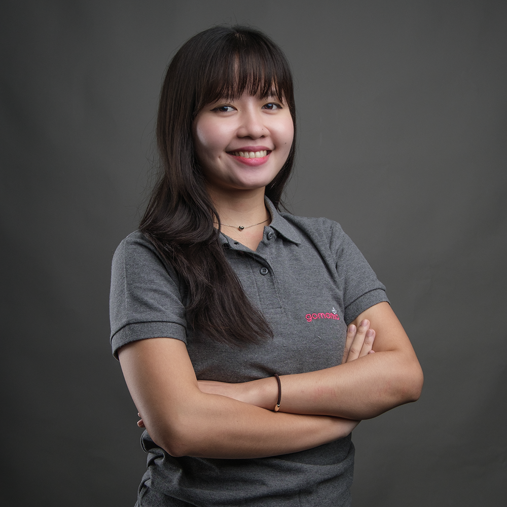 A young woman with long dark hair, wearing a gray polo shirt with pink embroidered text, stands with arms crossed and smiling at the camera against a gray background.