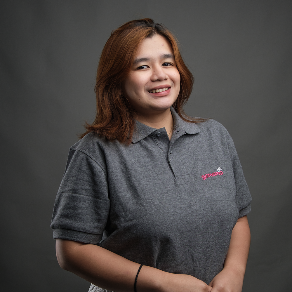 A young woman with shoulder-length brown hair smiling at the camera, wearing a gray polo shirt with a small pink logo on the left chest, standing against a dark gray background.