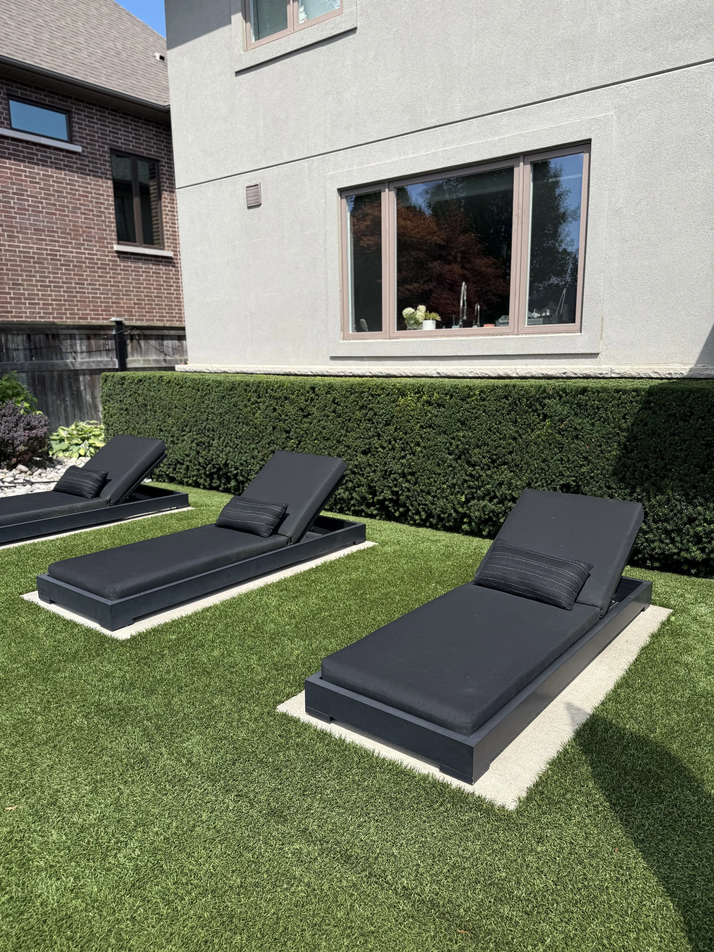 Backyard with three black lounge chairs on green grass, manicured hedge, and a modern house with a large window reflecting trees.