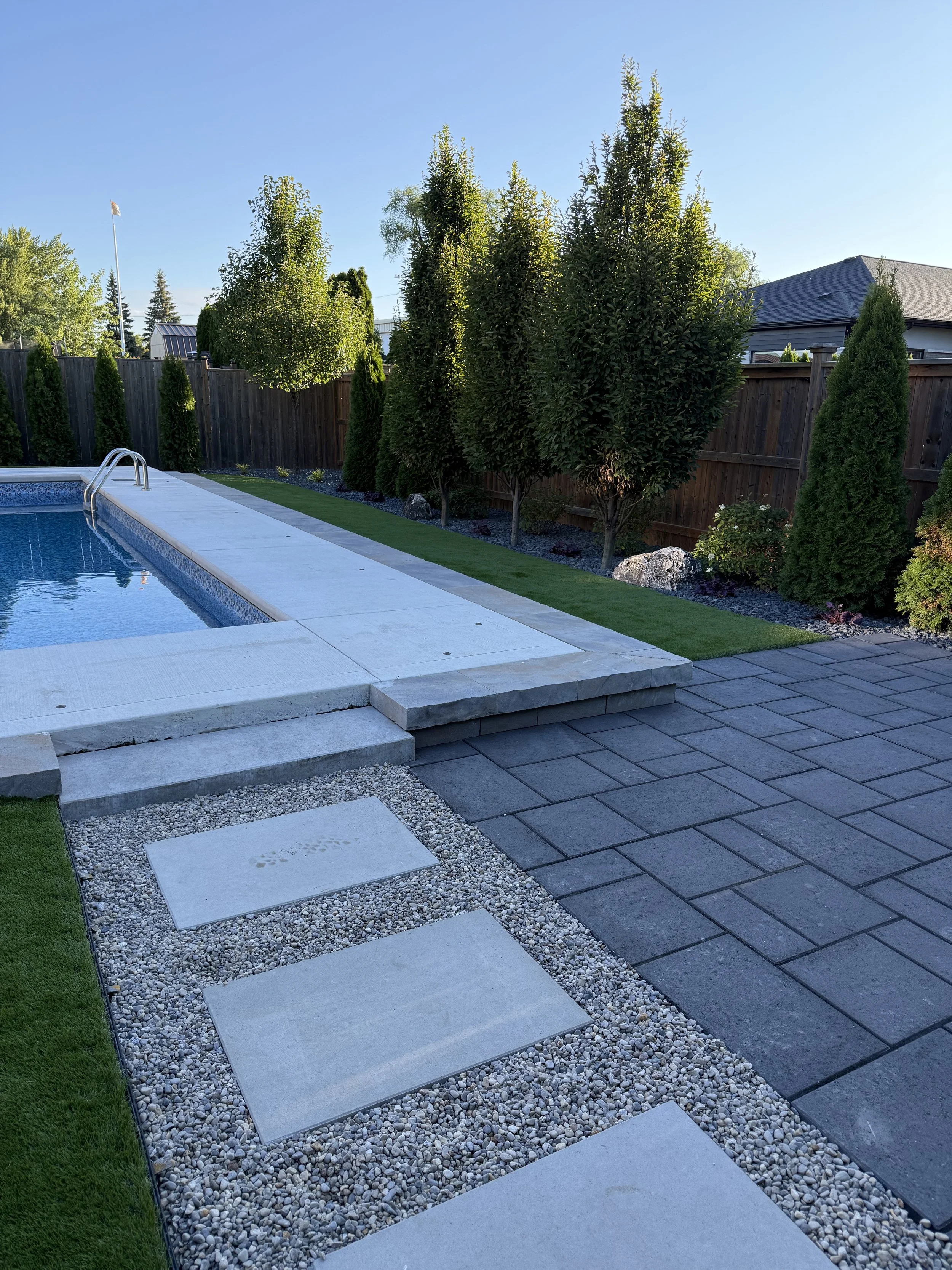 A backyard with a swimming pool, grass, a wooden fence, and various trees and shrubs, with a clear blue sky.