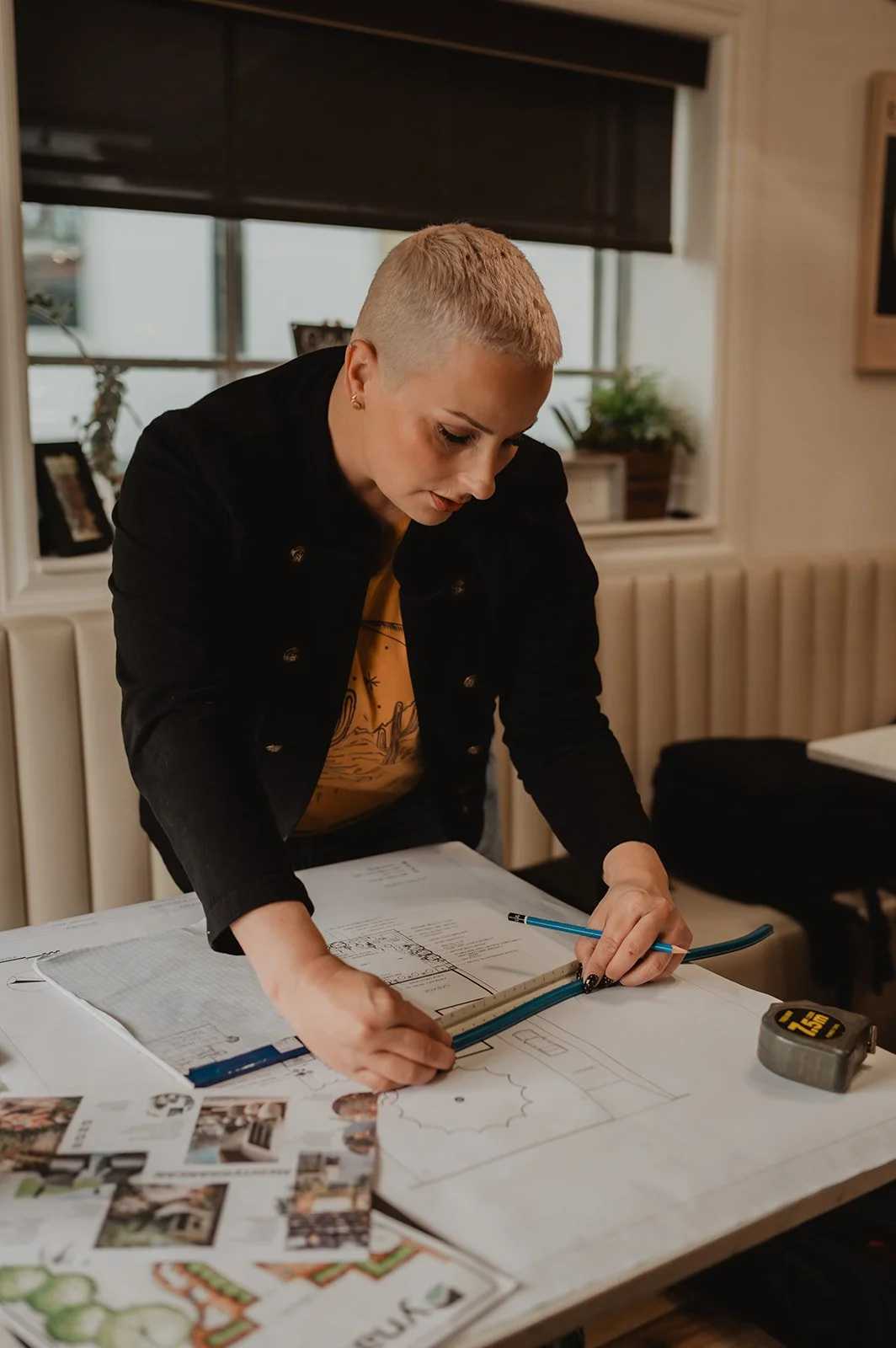 A woman with a buzz cut hairstyle working on architectural plans at a table, using a ruler and pencil, with photographs and drawings spread out on the table.