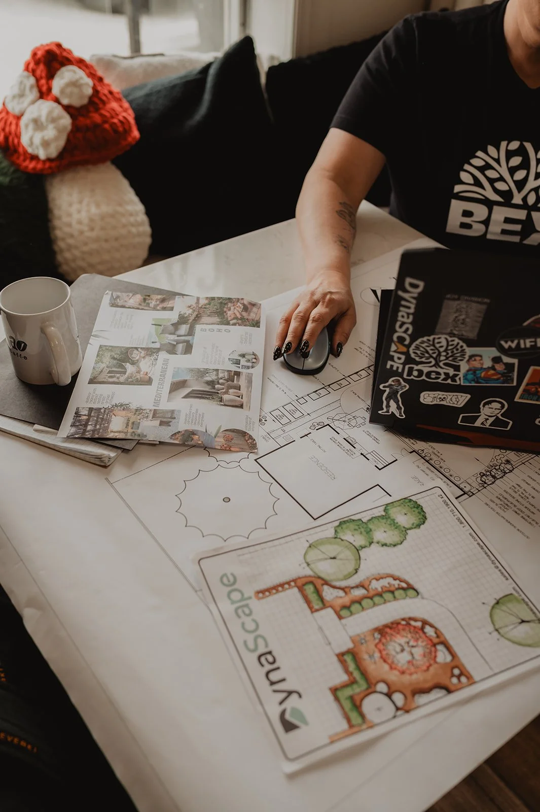 A person working at a desk with architectural plans, a brochure, a laptop covered in stickers, and a coffee mug.