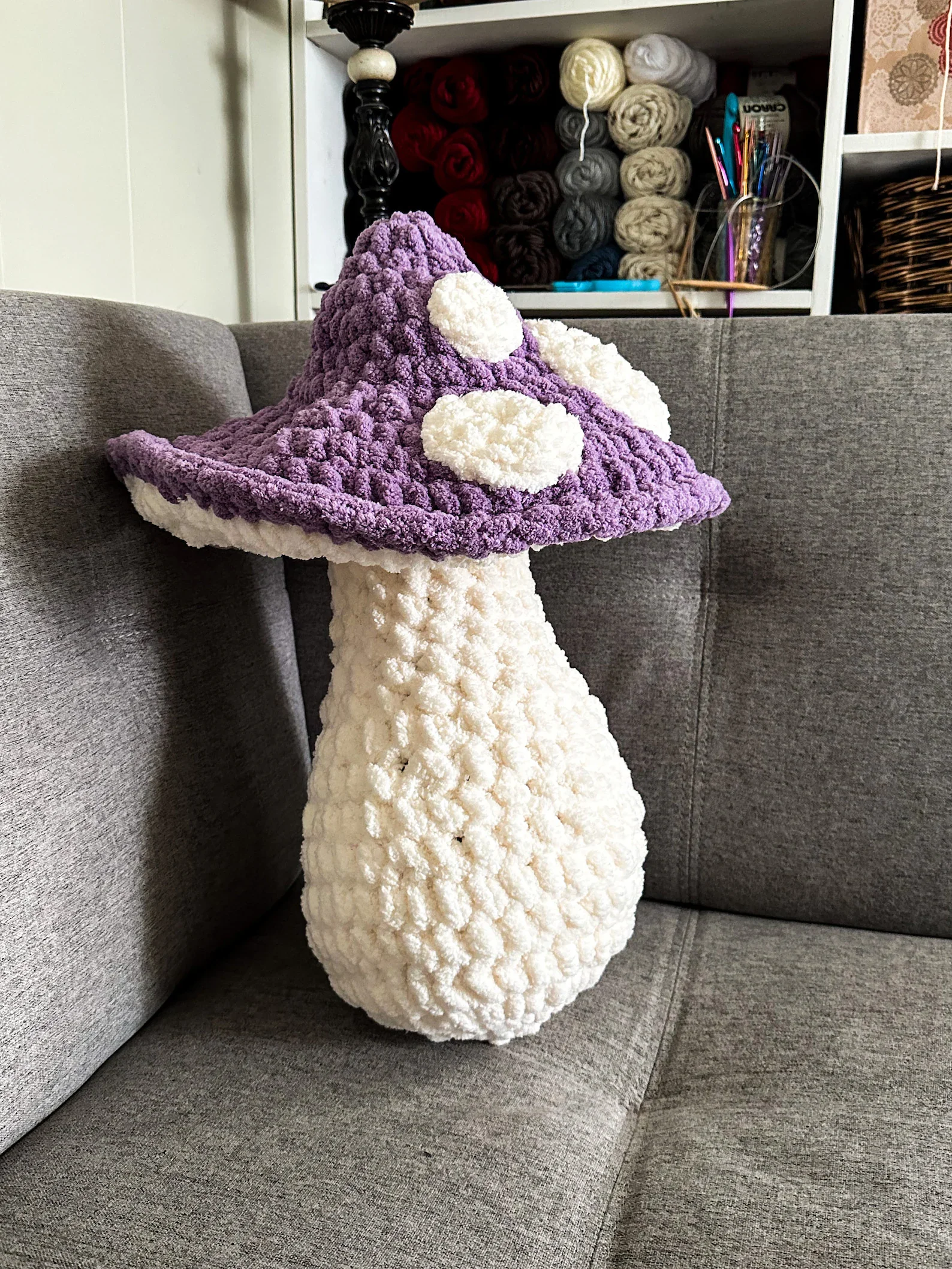 A plush mushroom-shaped pillow with a purple cap and white stem, placed on a gray sofa.