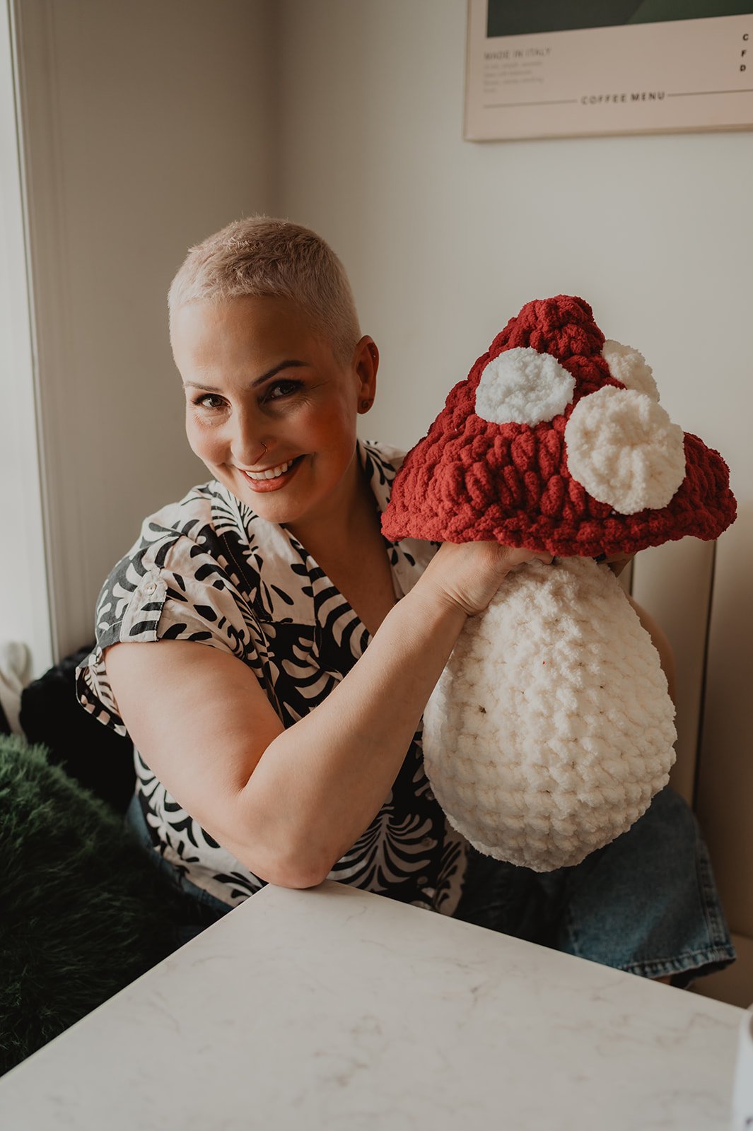 A woman with a short blonde hairstyle and a nose piercing holding a plush Christmas hat with a red body and white pompoms, smiling at the camera indoors.