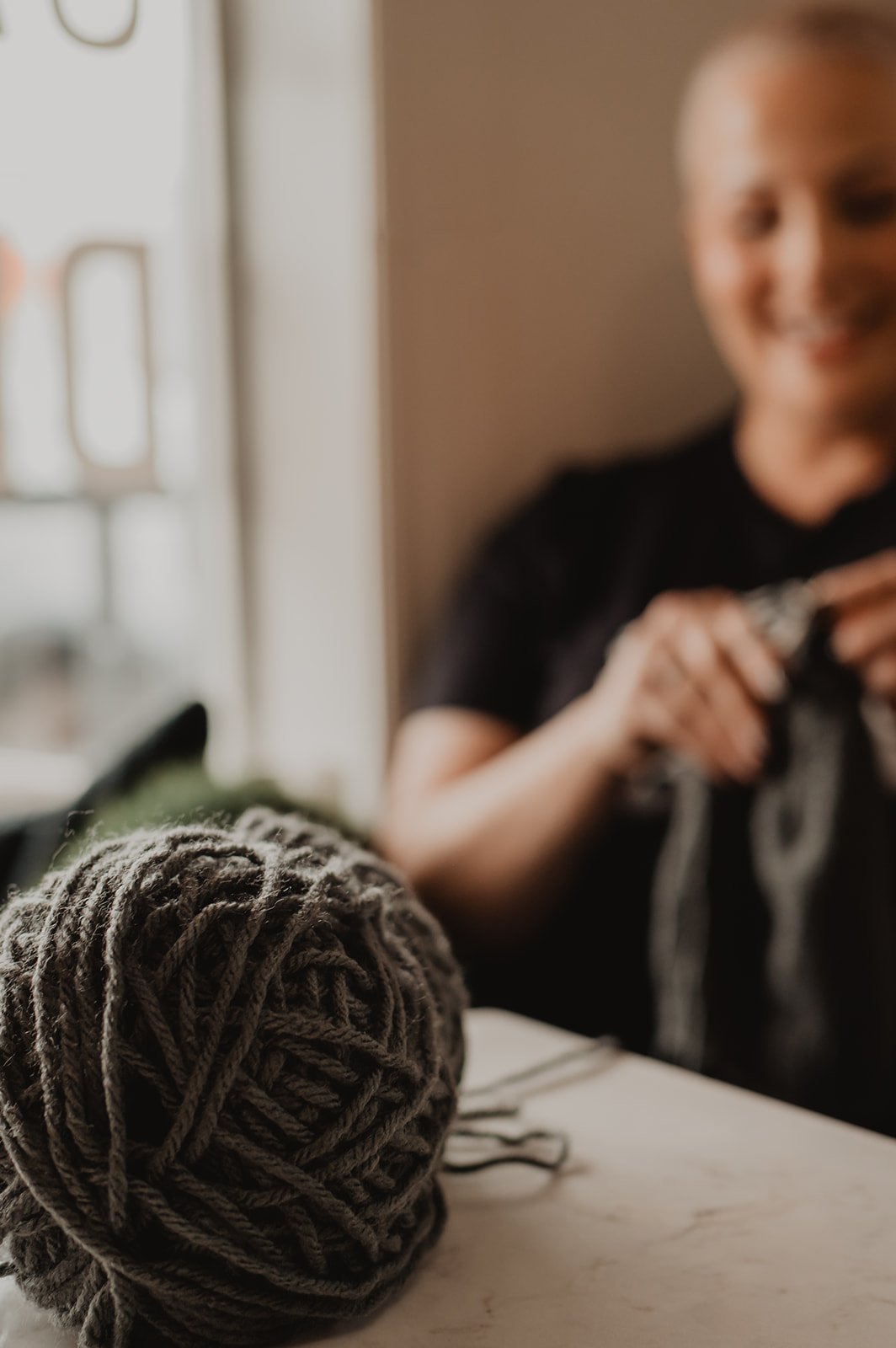 A ball of yarn in the foreground with a woman in the background holding knitting needles and smiling.