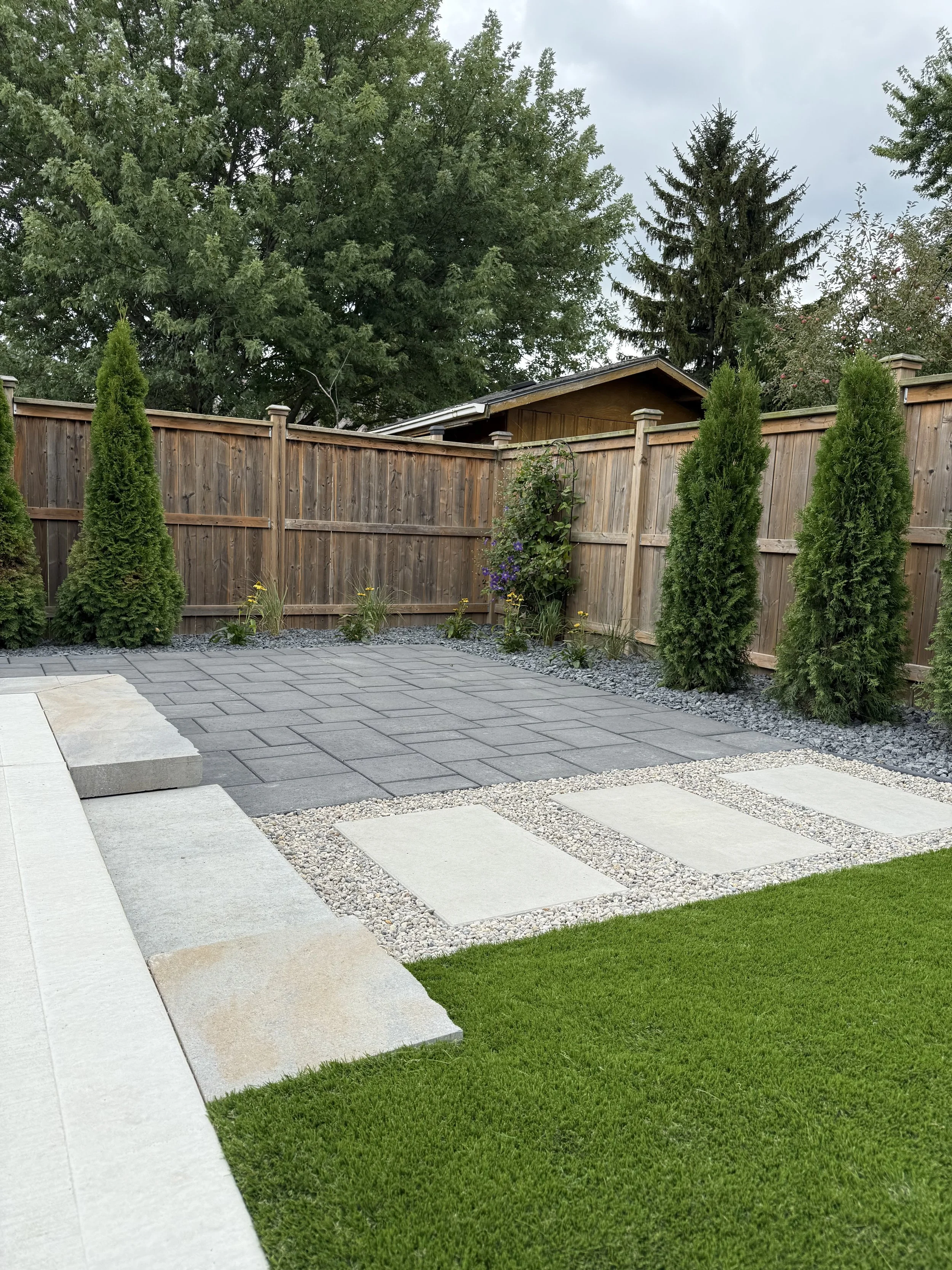 A backyard with a wooden privacy fence, trees, and a paved area with grass. The paved area has large concrete pavers and gravel sections. There are small shrubs and plants near the fence, with a shed roof peeking over the fence.
