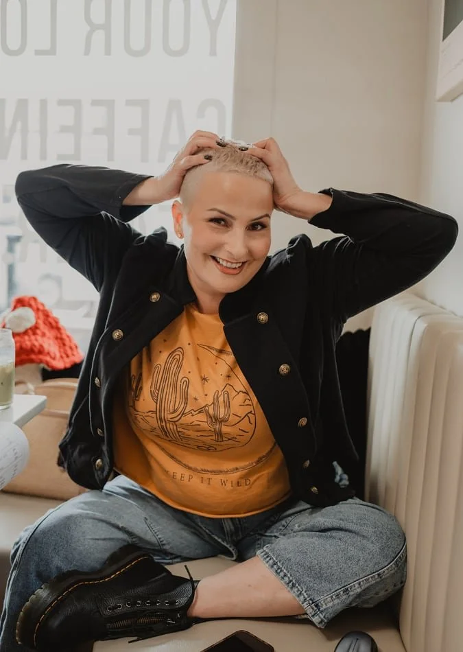 A smiling woman with a short blonde buzz cut, sitting cross-legged and holding her head with both hands, wearing a black jacket and a yellow graphic T-shirt, sitting on a cushioned bench inside a cafe or restaurant.