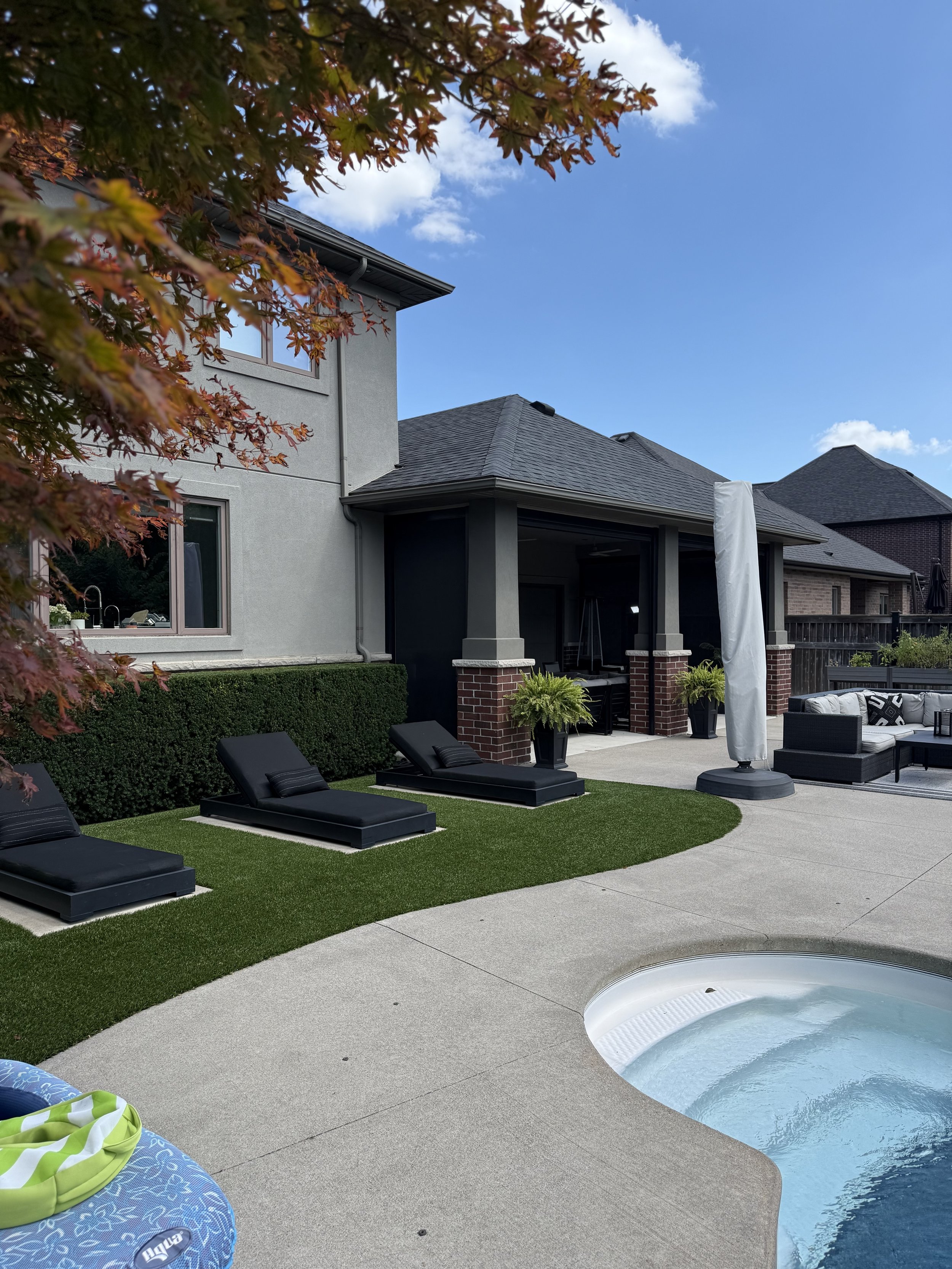 Backyard scene with a swimming pool, black lounge chairs, potted plants, and a house with a porch, under a partly cloudy sky.