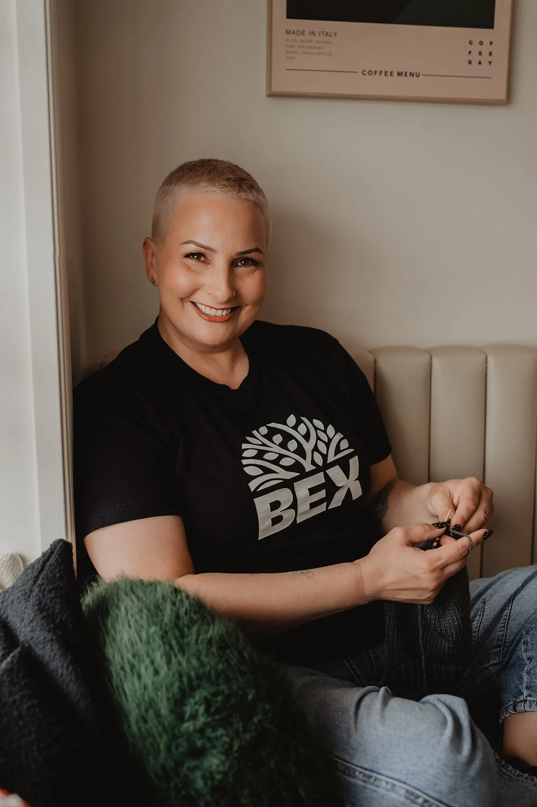 A woman with short blonde hair and a nose ring, smiling while sitting in a cozy indoor space. She is wearing a black t-shirt with a tree and the word 'BEX' on it, and holding a phone in her hands.