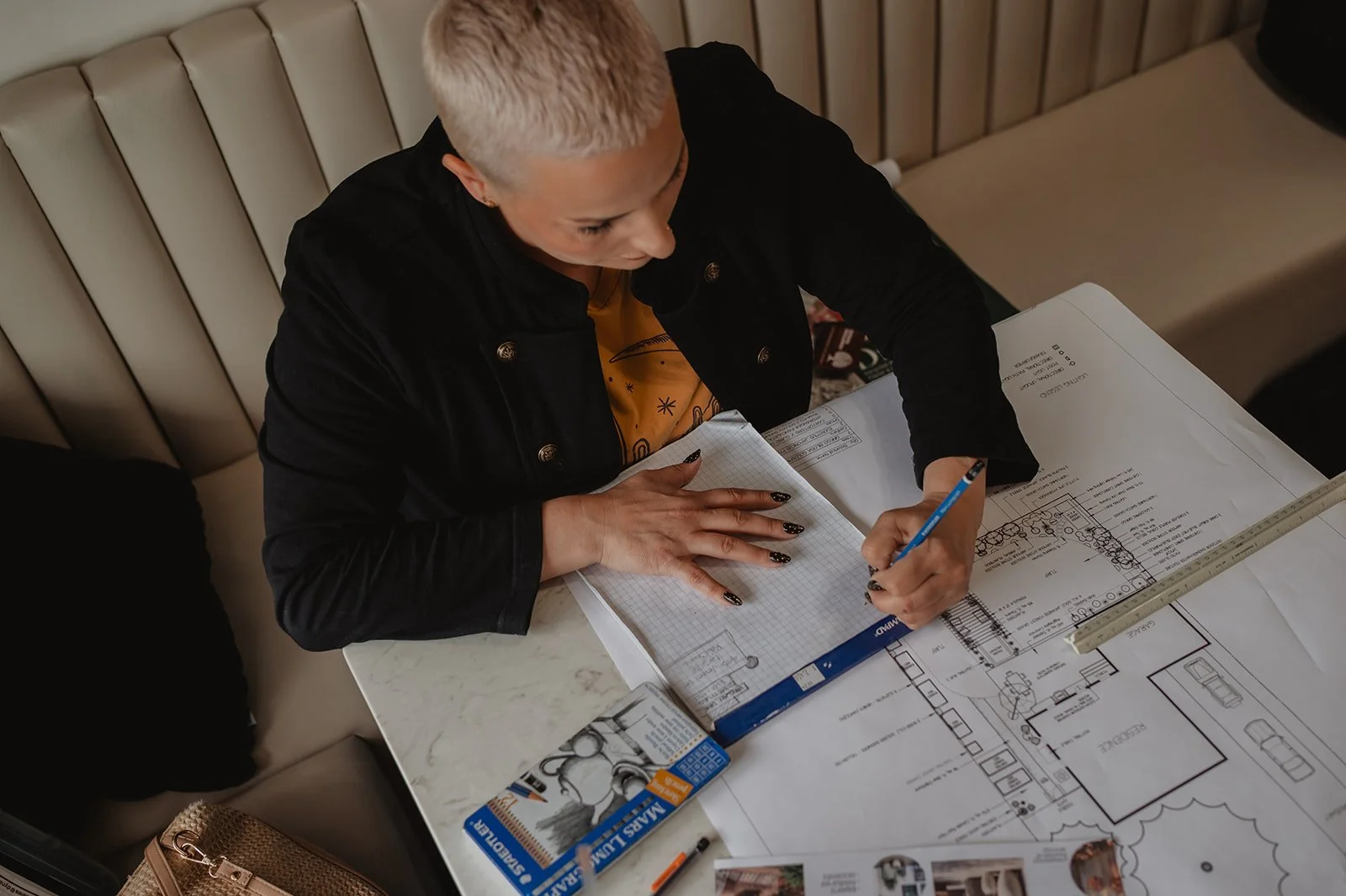 A person with short blonde hair and black nail polish is working on architectural plans and sketches at a table, with a ruler and a notebook nearby.