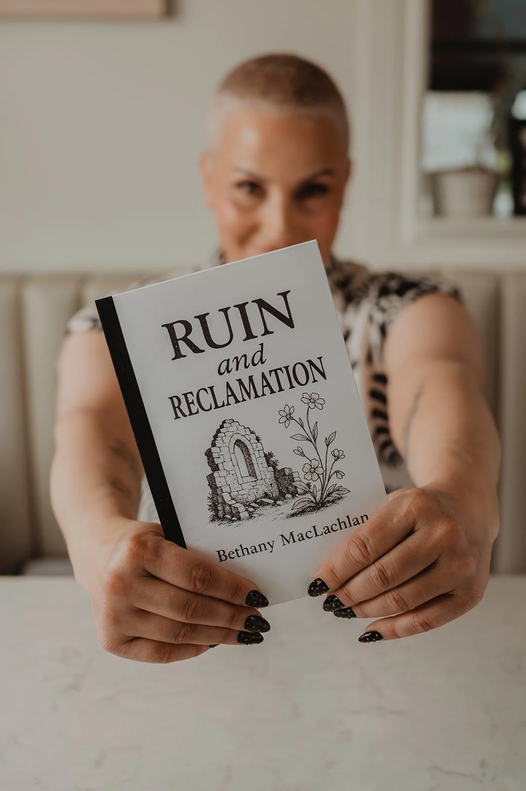 Woman with short hair holding a book titled 'Ruin and Reclamation' by Bethany MacLachlan, with a blurred background.
