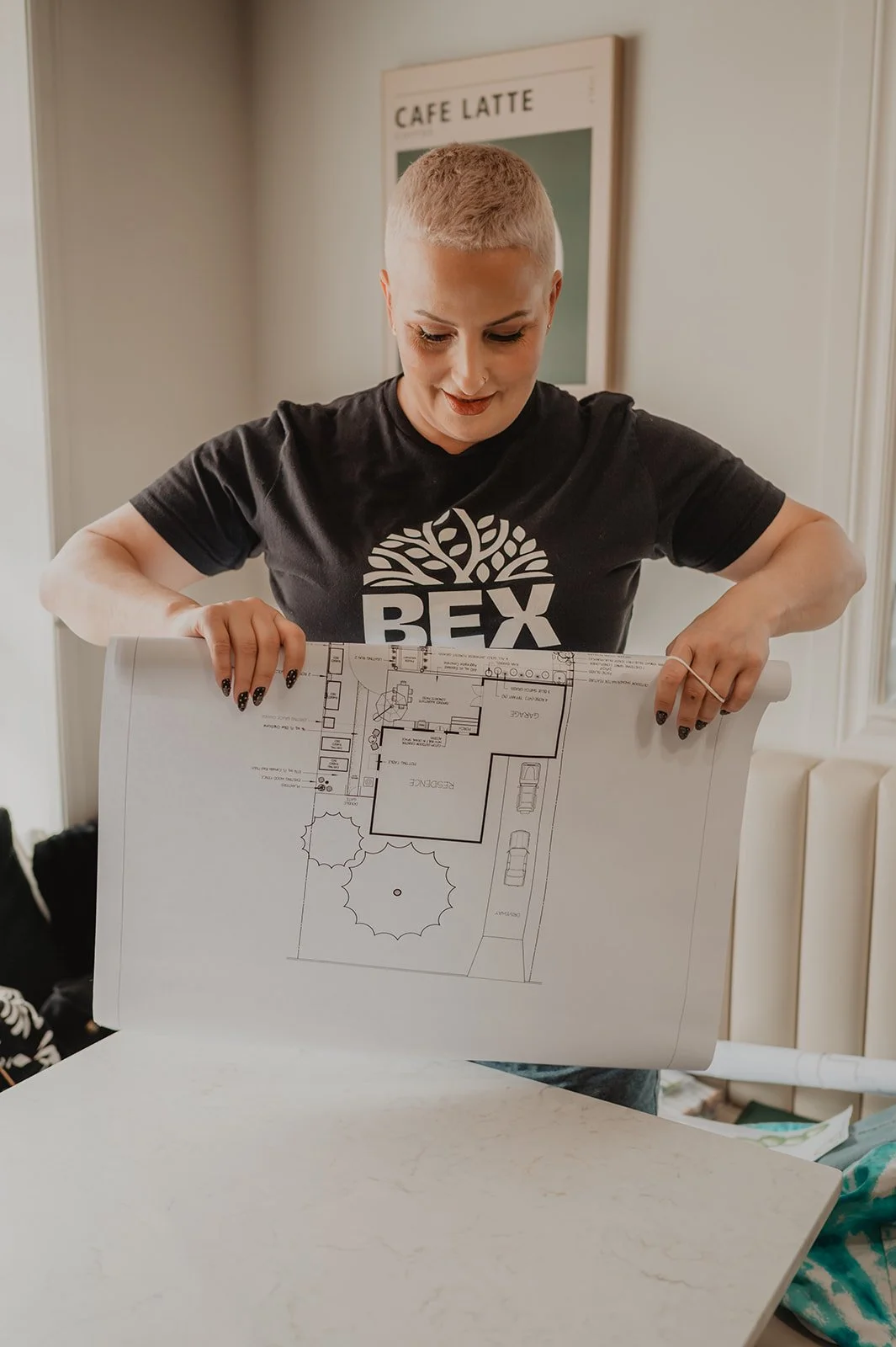A woman with short blond hair and black nail polish holds a large architectural blueprint, looking down at it. She wears a black T-shirt with a logo that says 'BEX' and a tree design. Behind her is a wall with a 'Cafe Latte' sign.