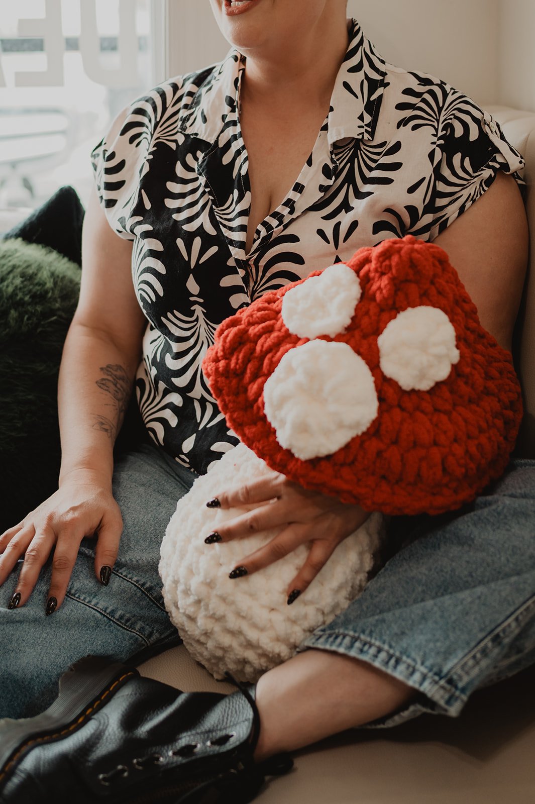 Person sitting on a couch holding a large, plush, red and white object resembling a mushroom with white spots, wearing a black and white patterned shirt and jeans.