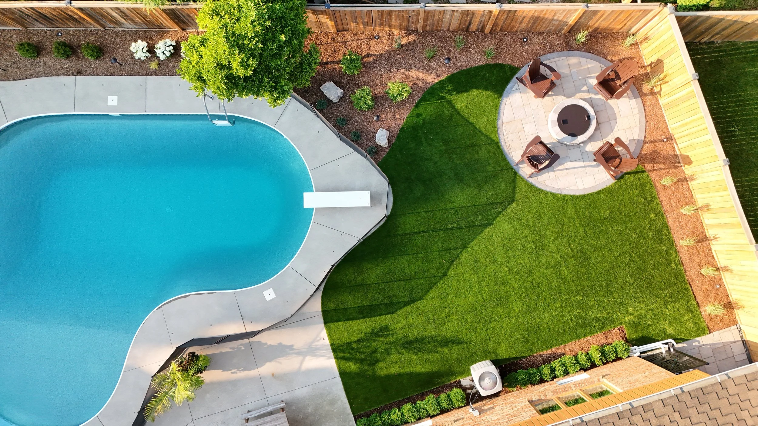 A backyard with a curved swimming pool, a small grassy area, a patio with four Adirondack chairs around a firepit, and a wooden fence with landscaping along the fence line.