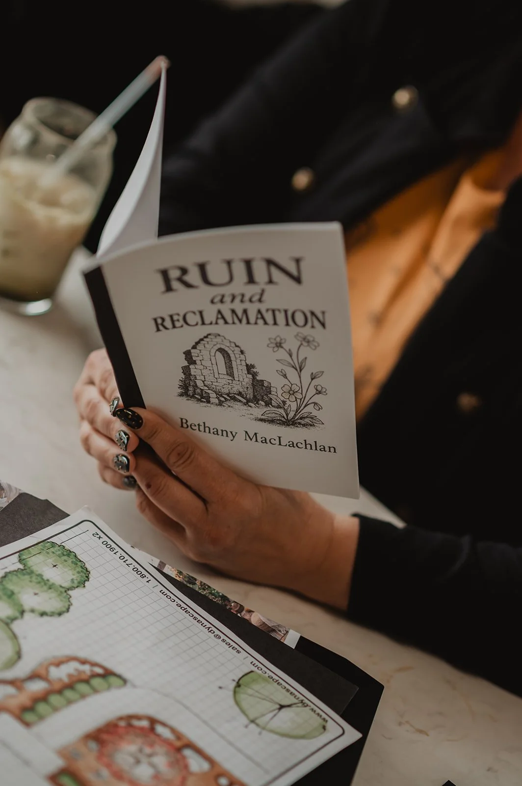 A person sitting at a table reading a book titled 'Ruin and Reclamation' by Bethany MacLachlan. The person has decorative black and silver nails, and there is a glass of iced coffee and a sheet of paper with sketches of trees and landscape designs on