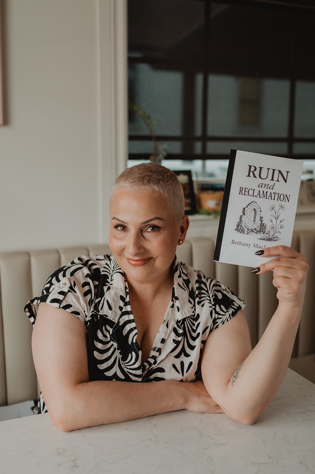 A woman with short blonde hair, smiling, holding a book titled 'Ruin and Reclamation' by Bethany MacL. She is sitting at a table in a restaurant or cafe, wearing a black and white patterned top.