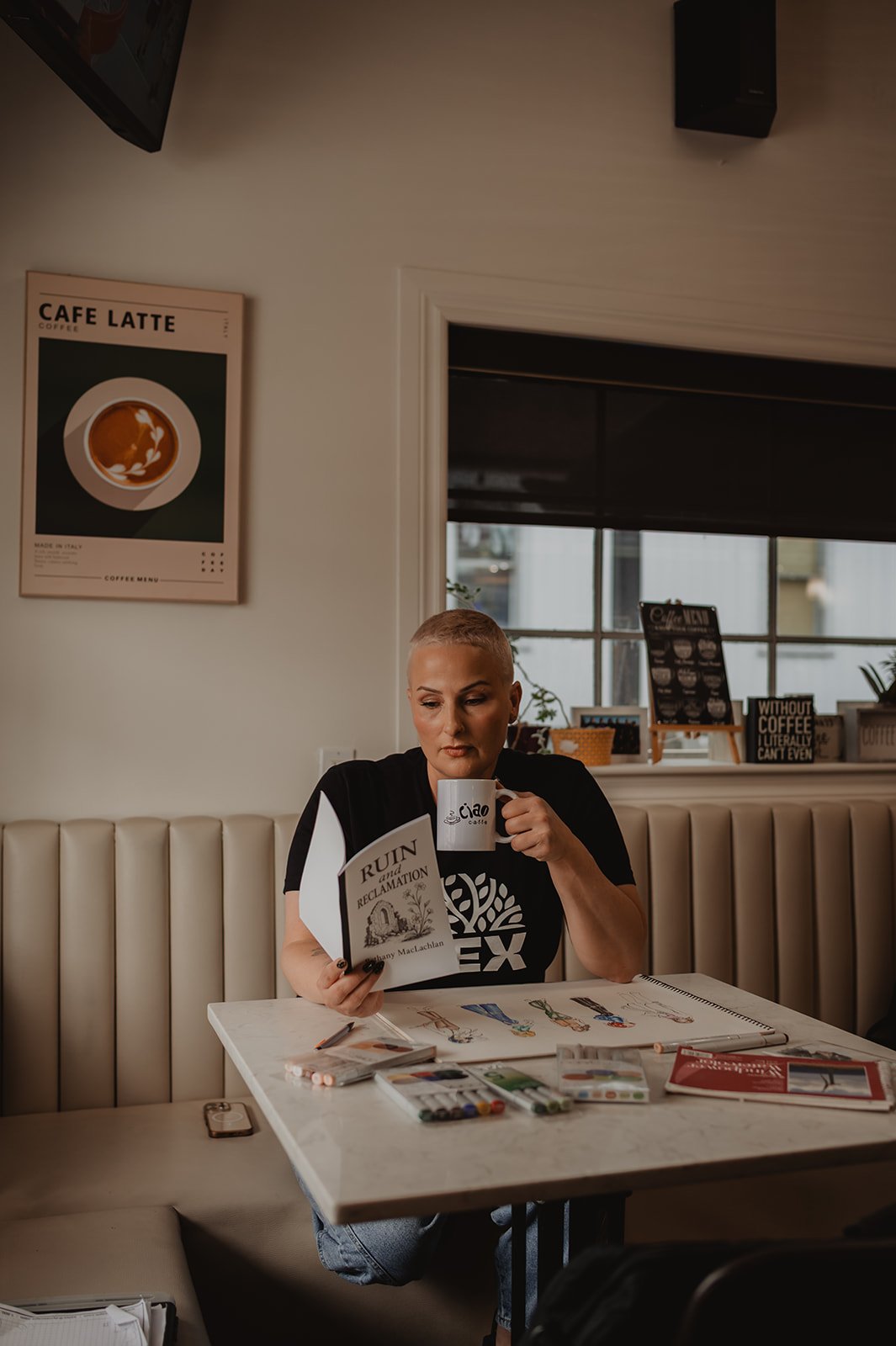 Woman with short blonde hair sitting at a table in a coffee shop, reading a book titled 'Ruin and Reclamation', holding a white mug with the cafe's logo, surrounded by art supplies and sketchbooks.