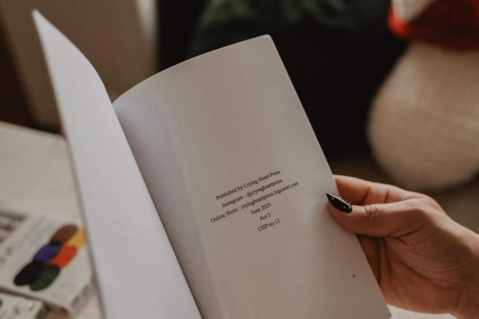 A person holding an open book, showing publication details on the right page, including publisher, Instagram handle, website, and date; blurred background with a painted object on a table.