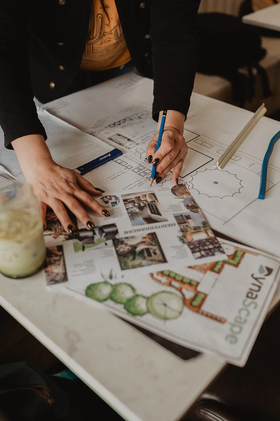 Person working on architectural plans and a printed brochure with photographs of home interiors and exteriors, on a white table.