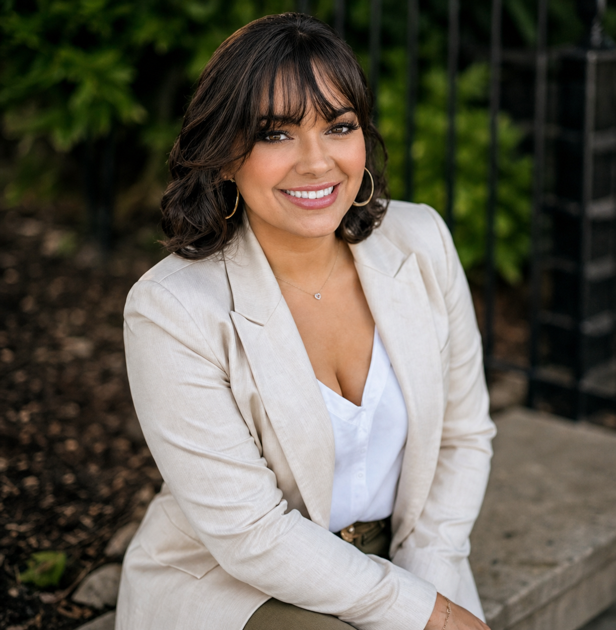 A woman with dark, shoulder-length hair and bangs, smiling while sitting outdoors in front of greenery, wearing a light-colored blazer, white top, gold hoop earrings, and a necklace.