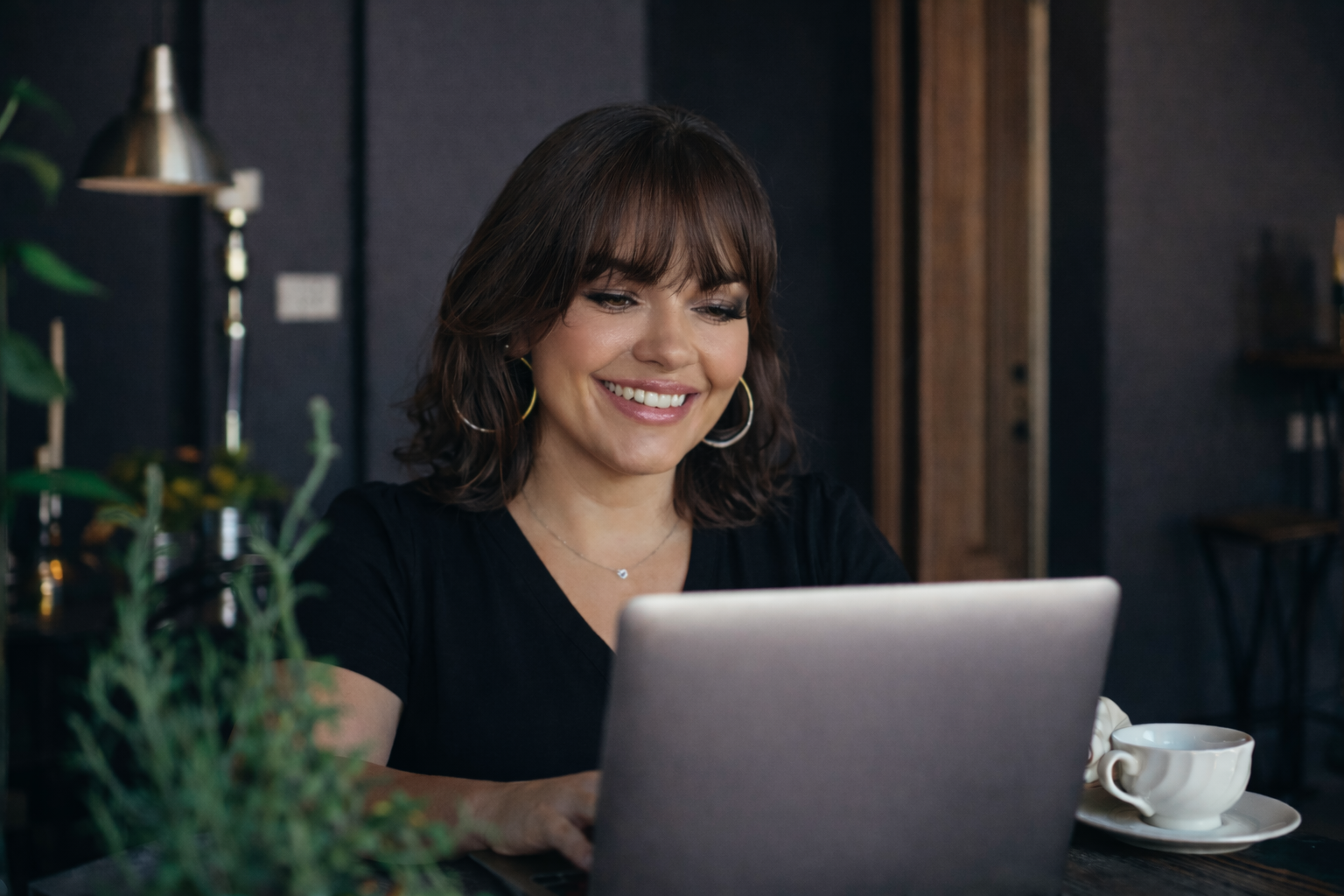 A woman with shoulder-length brown hair and hoop earrings sits at a table, smiling while working on a laptop, with a cup of coffee nearby in a cozy cafe setting.