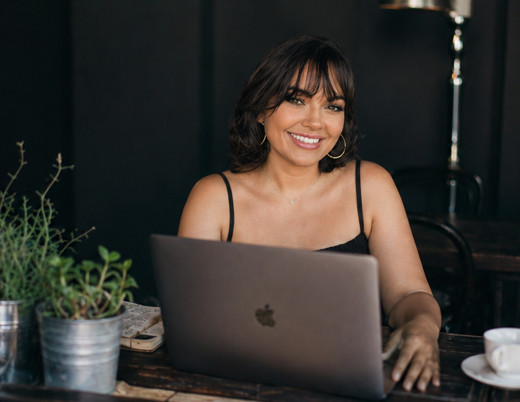 A woman with shoulder-length dark hair and hoop earrings smiling while working on a laptop at a wooden table, with potted plants and a white cup nearby.