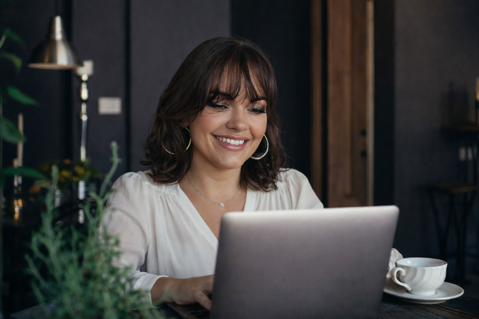 A woman with shoulder-length brown hair, wearing a white blouse and hoop earrings, is smiling while using a laptop in a cozy, modern cafe setting with dark walls, a pendant light, and a table setting with a white coffee cup.