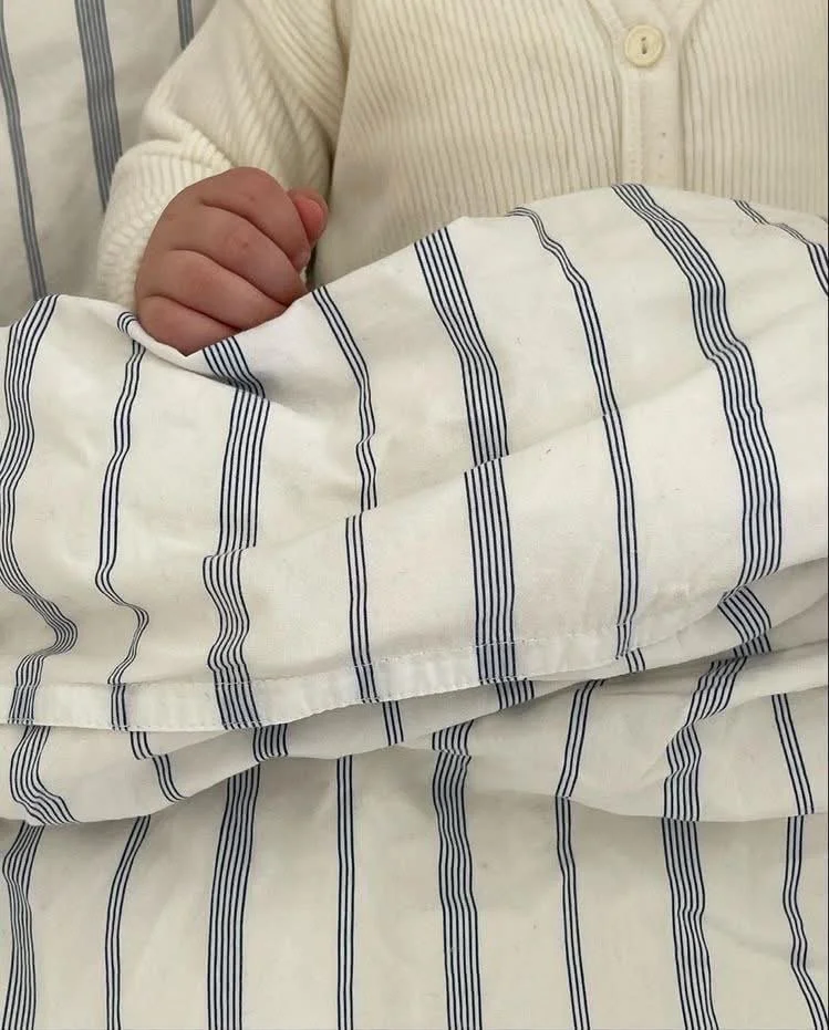 Billedet viser en person i en hvid cardigan, der holder en baby, der er indpakket i en stofble med blå striber.