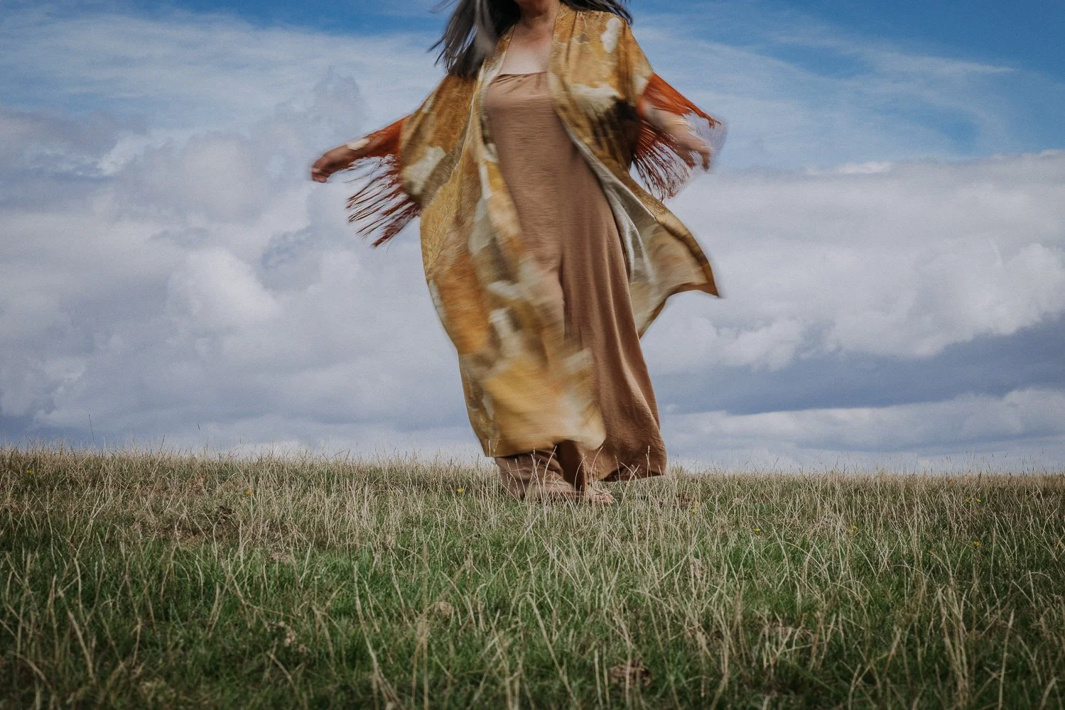 A woman in a flowing dress and a patterned shawl standing barefoot on a grassy field under a cloudy sky.