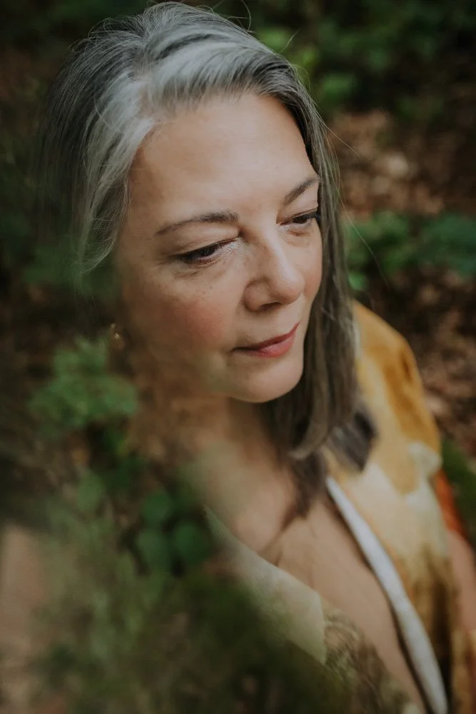 Close-up of a woman with gray and brown hair, looking down, outdoors in a natural setting with greenery and soil background.