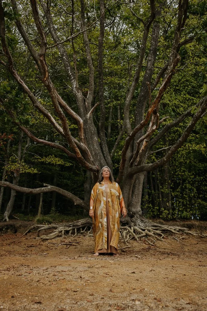 A woman dressed in a long beige and gold patterned robe stands barefoot in front of a large, ancient tree with extensive roots in a forest setting.