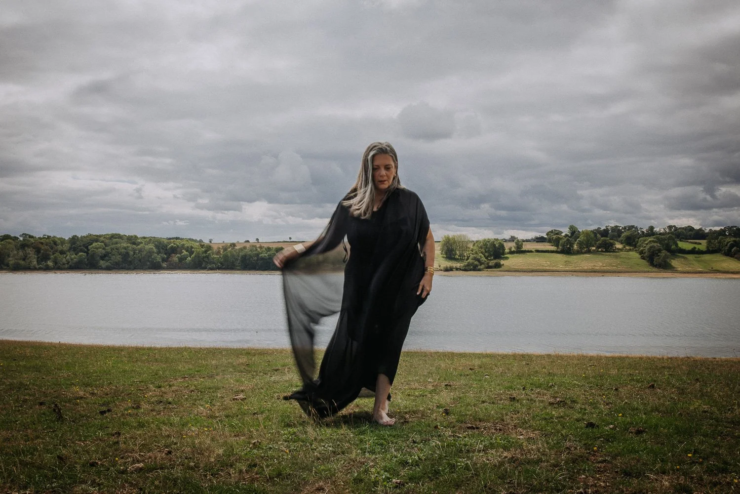 A woman in black clothing stands barefoot on grass near a body of water on an overcast day, holding a sheer black shawl.