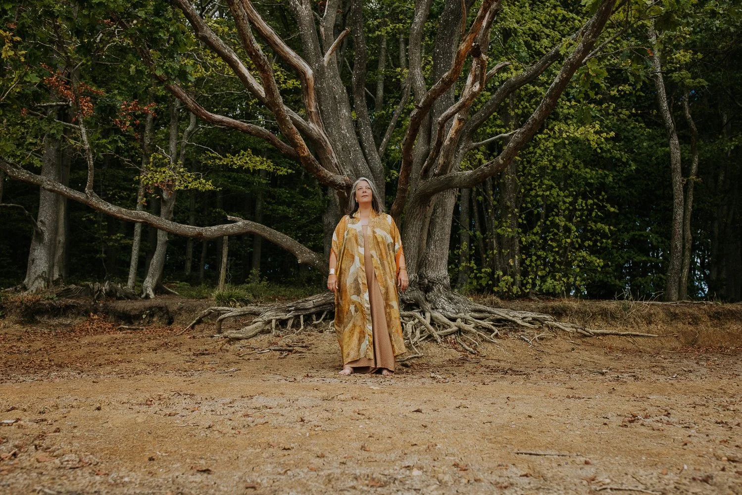 A woman standing barefoot on a dirt ground in front of a large tree in a forest, looking upwards with her hands at her sides, wearing a long patterned coat over a tan dress.