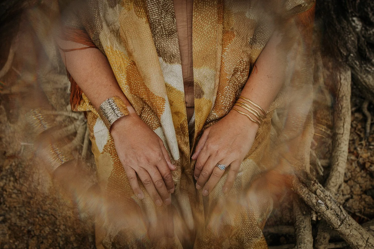 Close-up of a person's hands with jewelry, sitting outdoors on dirt and tree roots, wearing a patterned yellow and brown garment.