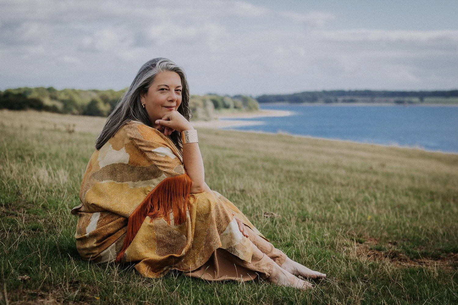 A woman with gray hair sitting on grass near a body of water, wearing a patterned shawl with fringe, smiling softly, facing slightly to the camera, with a distant shoreline and cloudy sky in the background.
