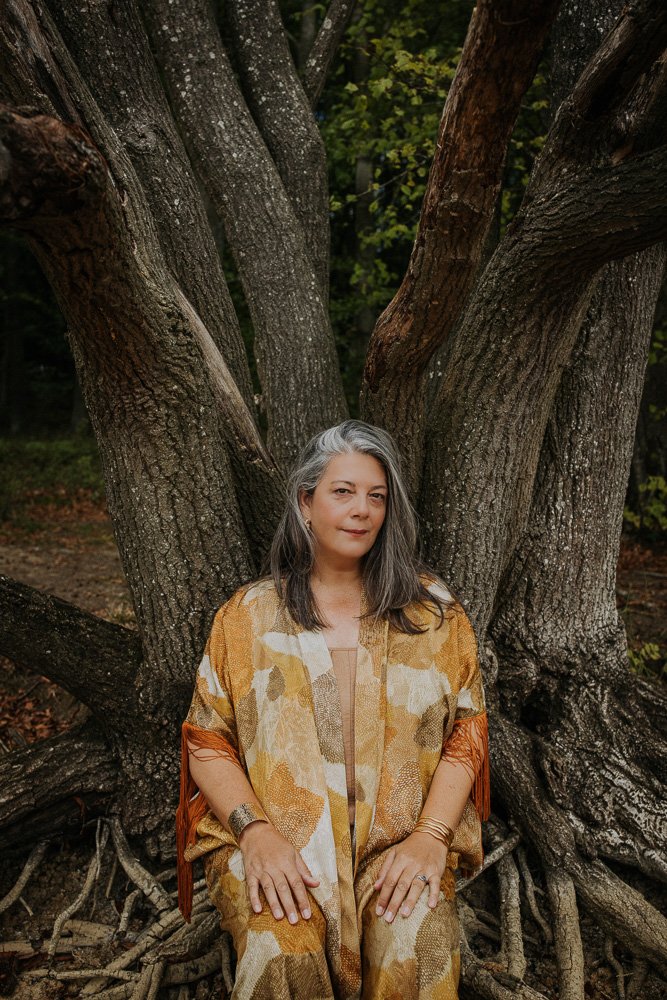 A woman with gray hair sitting against the roots of a large, gnarled tree in an outdoor setting with green foliage.