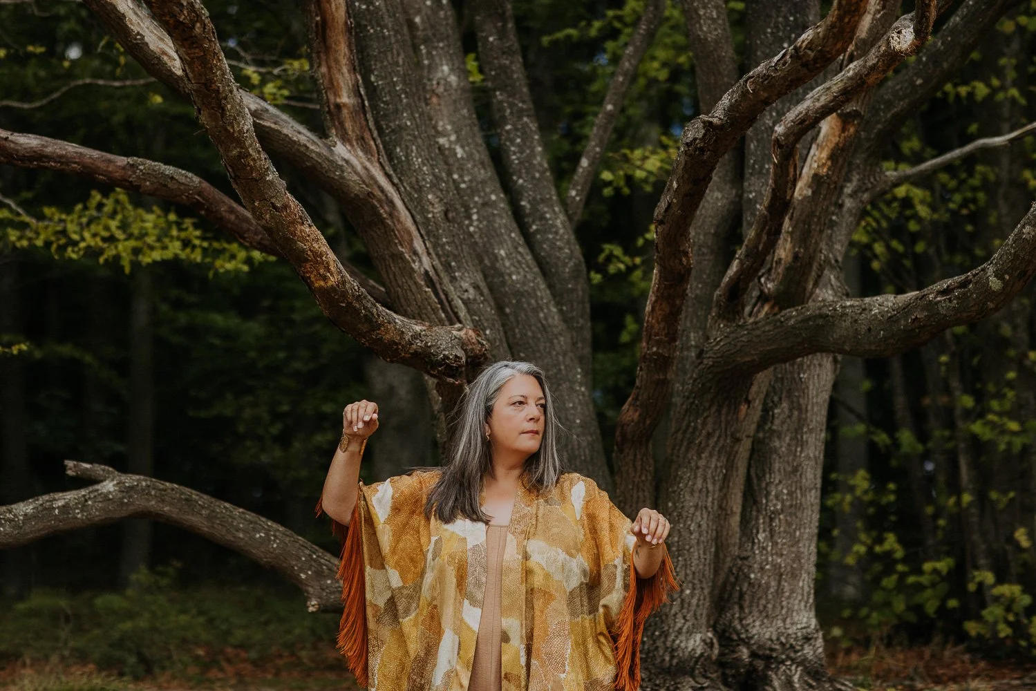 A woman with long gray hair standing outdoors in front of a large, leafless tree with multiple branches. She is wearing a patterned shawl with tassels and has her arms raised in a flexed position.