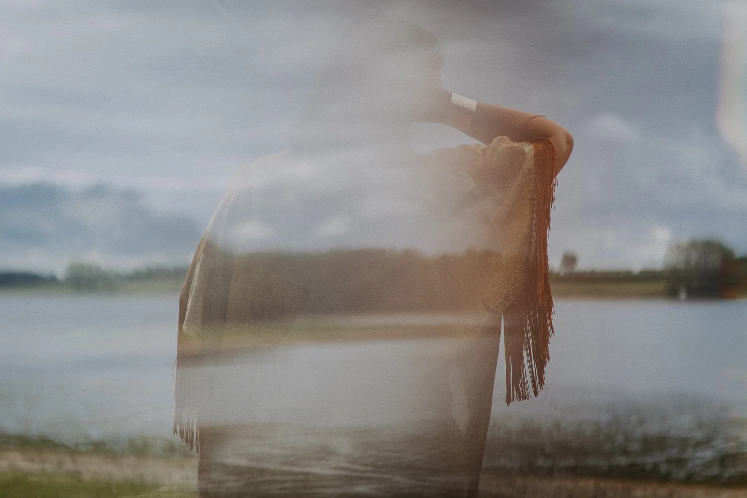 A person standing outdoors near a body of water, viewed through a transparent surface that reflects the sky and landscape, creating a layered double exposure effect.