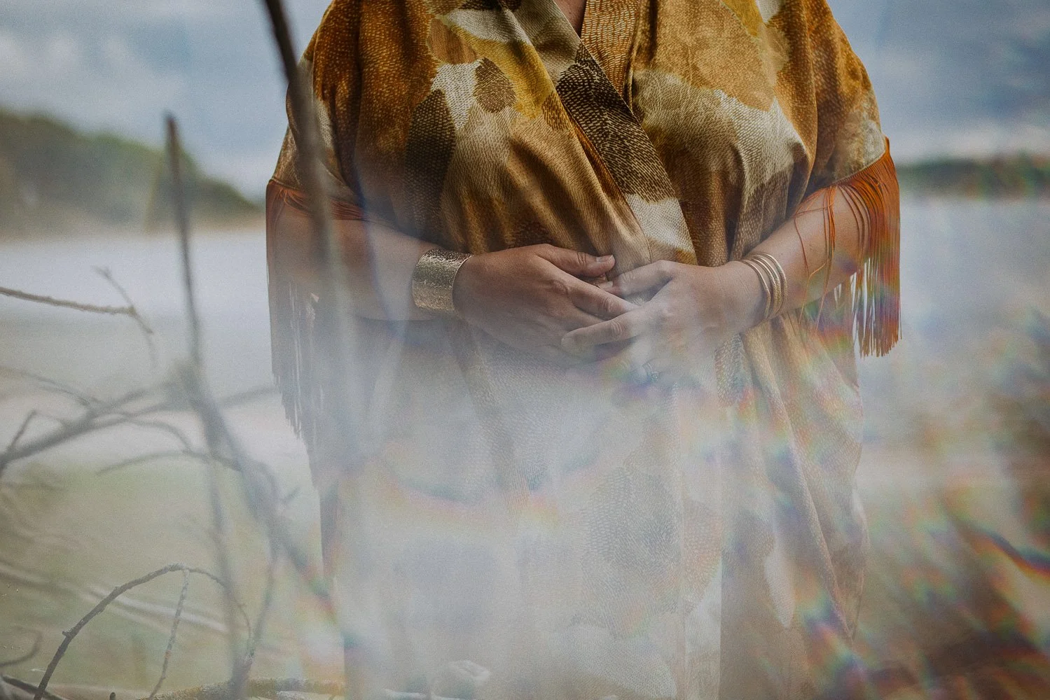 A woman wearing a fringed, multicolored shawl and silver jewelry, standing outdoors with a blurred background of water and trees, partially obscured by a reflection.